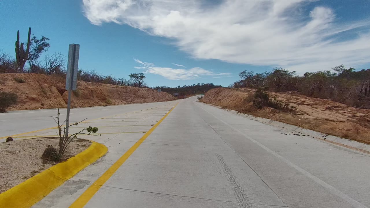 A paved road in East Cape Los Cabos, Baja California Sur passes red soil, sparse brush, a cactus, and a roadside post under bright clouds, with no visible people.