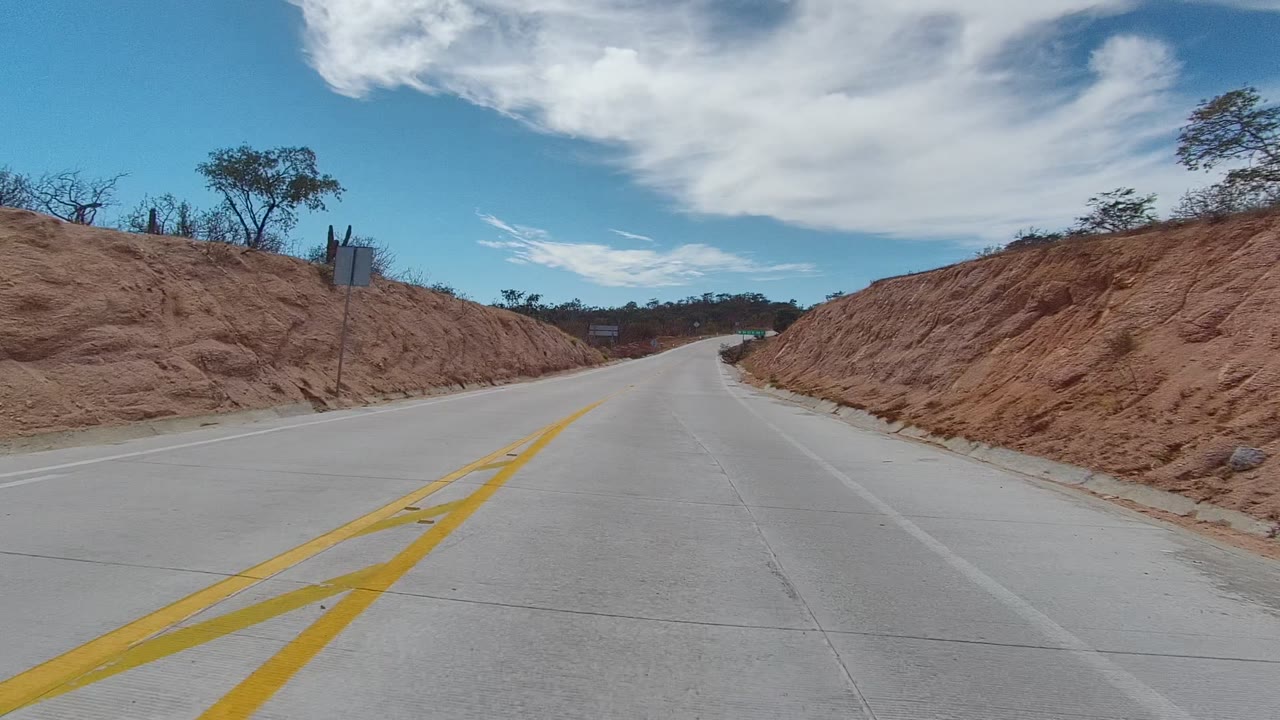 A curving paved road in East Cape Los Cabos passes a rounded red dirt mound, scattered brush, and a small roadside sign shape beneath a blue sky, with no visible people.