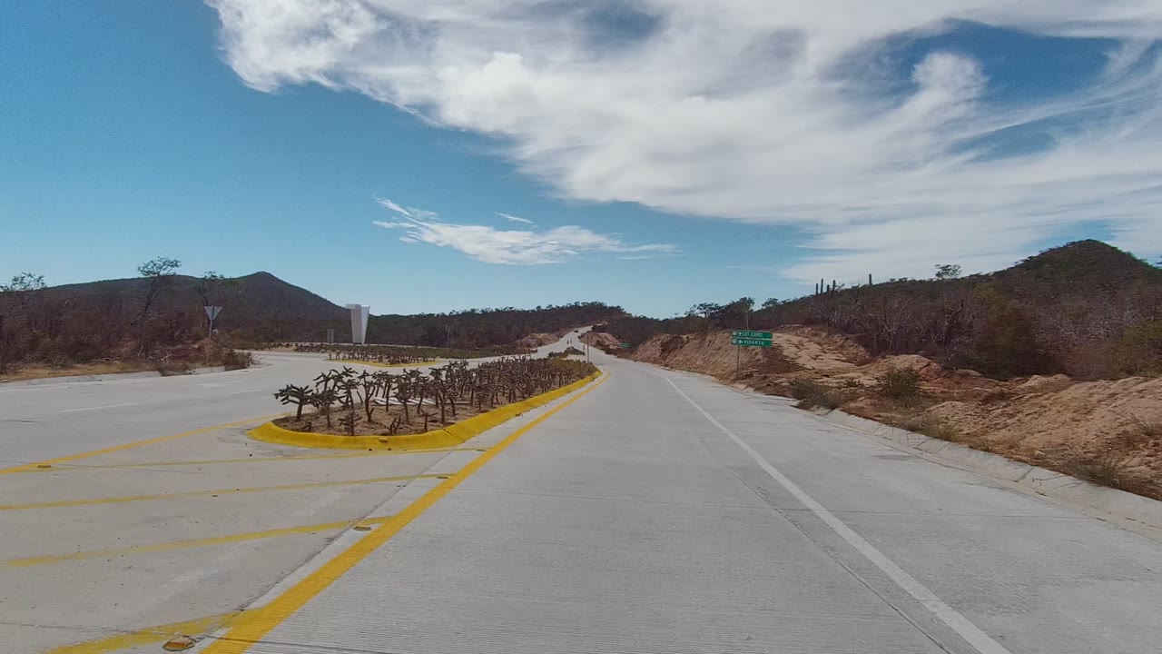 Opening view in East Cape Los Cabos, Mexico, looking along a concrete road toward a cactus roundabout, low hills, dry vegetation, and no visible people.