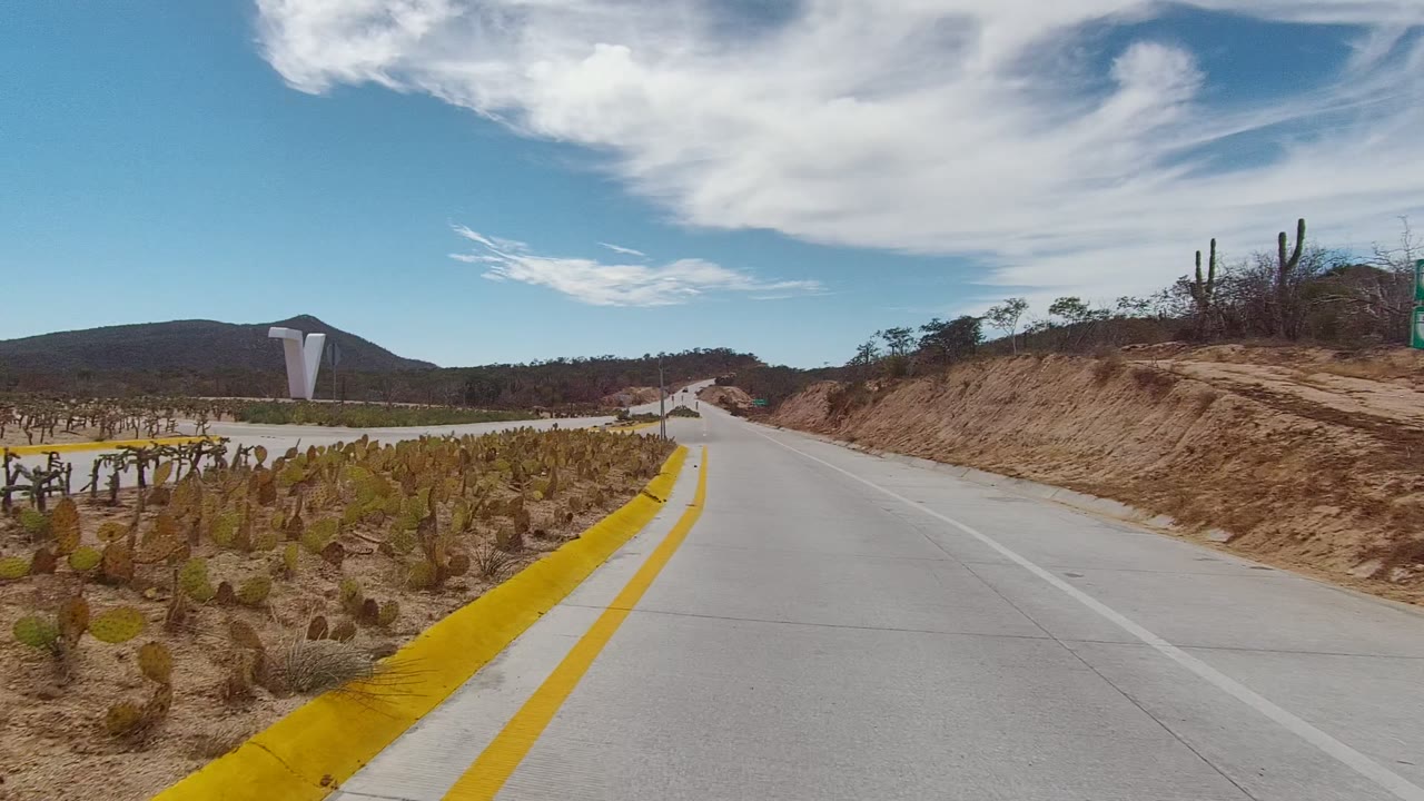 East Cape Los Cabos road view with a yellow-edged cactus median on the left, sandy roadside cut on the right, distant hills, and no visible people.