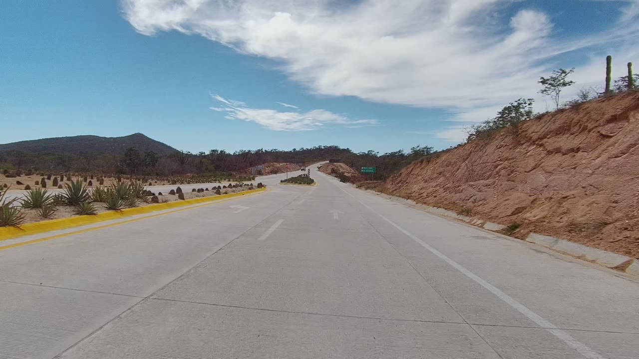 East Cape Los Cabos road view at the start of the clip, with a concrete lane, yellow curb, planted desert vegetation, distant low mountains, broad white clouds, and no visible people.
