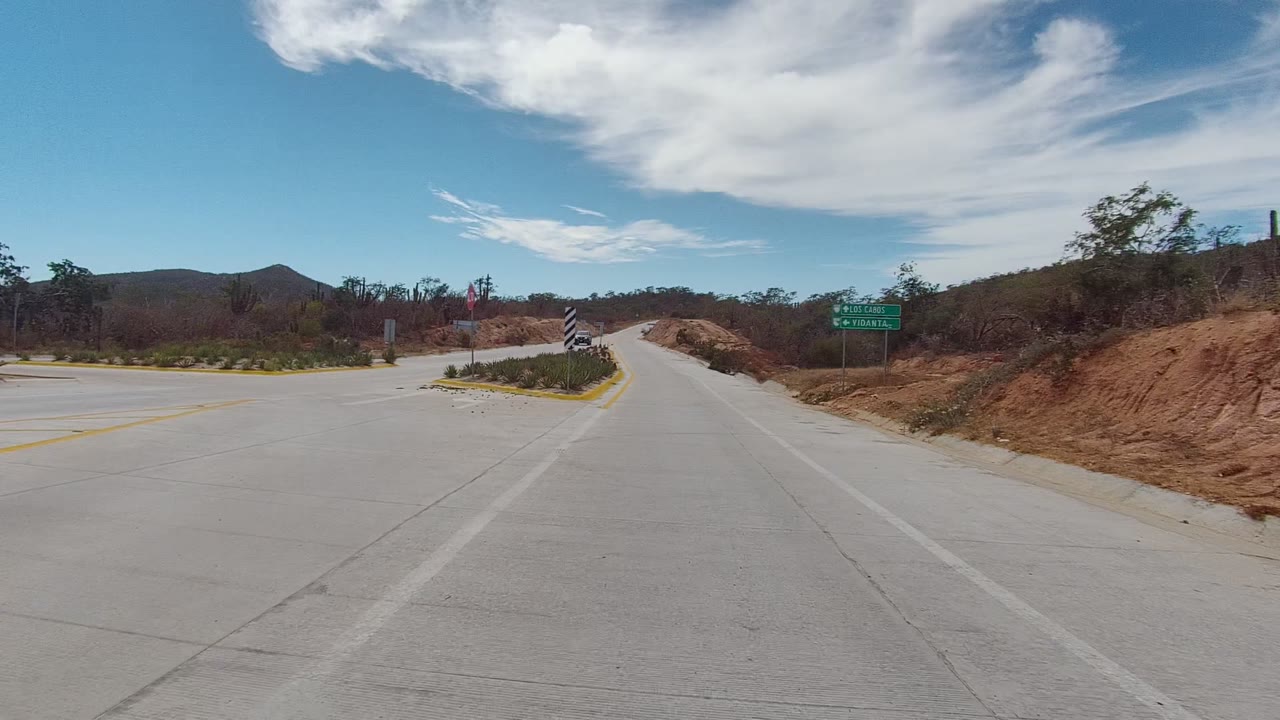 East Cape Los Cabos roadway view at two seconds, with a curving concrete lane, green directional signs, dry brush, reddish roadside earth, distant hills, high clouds, and no visible people.