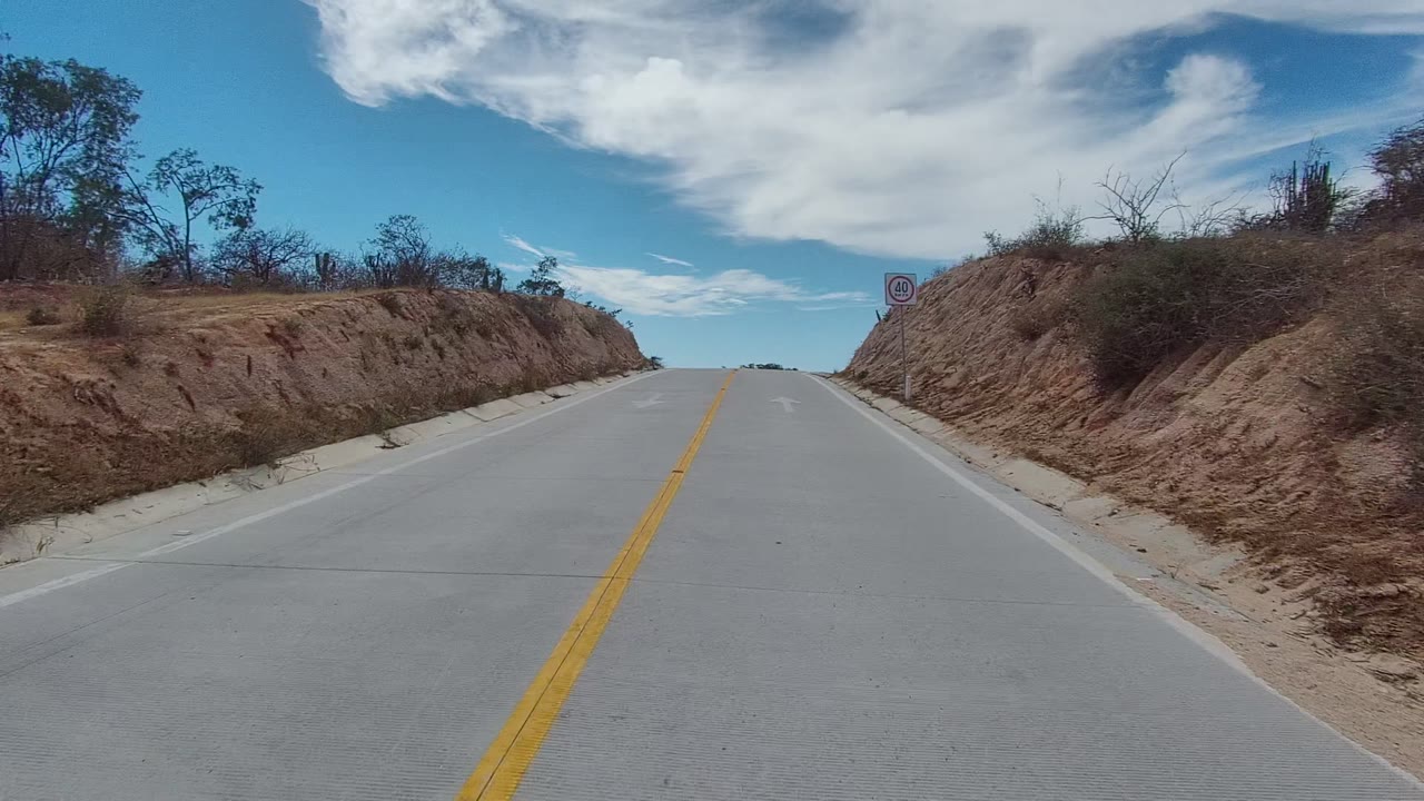 A two-lane road in East Cape Los Cabos, Baja California Sur, Mexico climbs between dry cut slopes with yellow center lines, sparse vegetation, white clouds, and no visible people.
