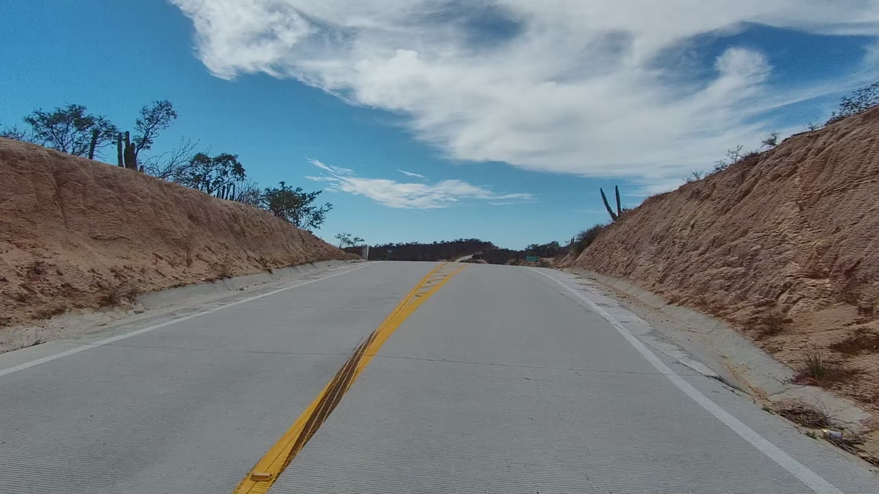 A curving section of road in East Cape Los Cabos passes between dry slopes, with double yellow markings in the foreground, scattered cactus and trees on the hillsides, and no visible people.