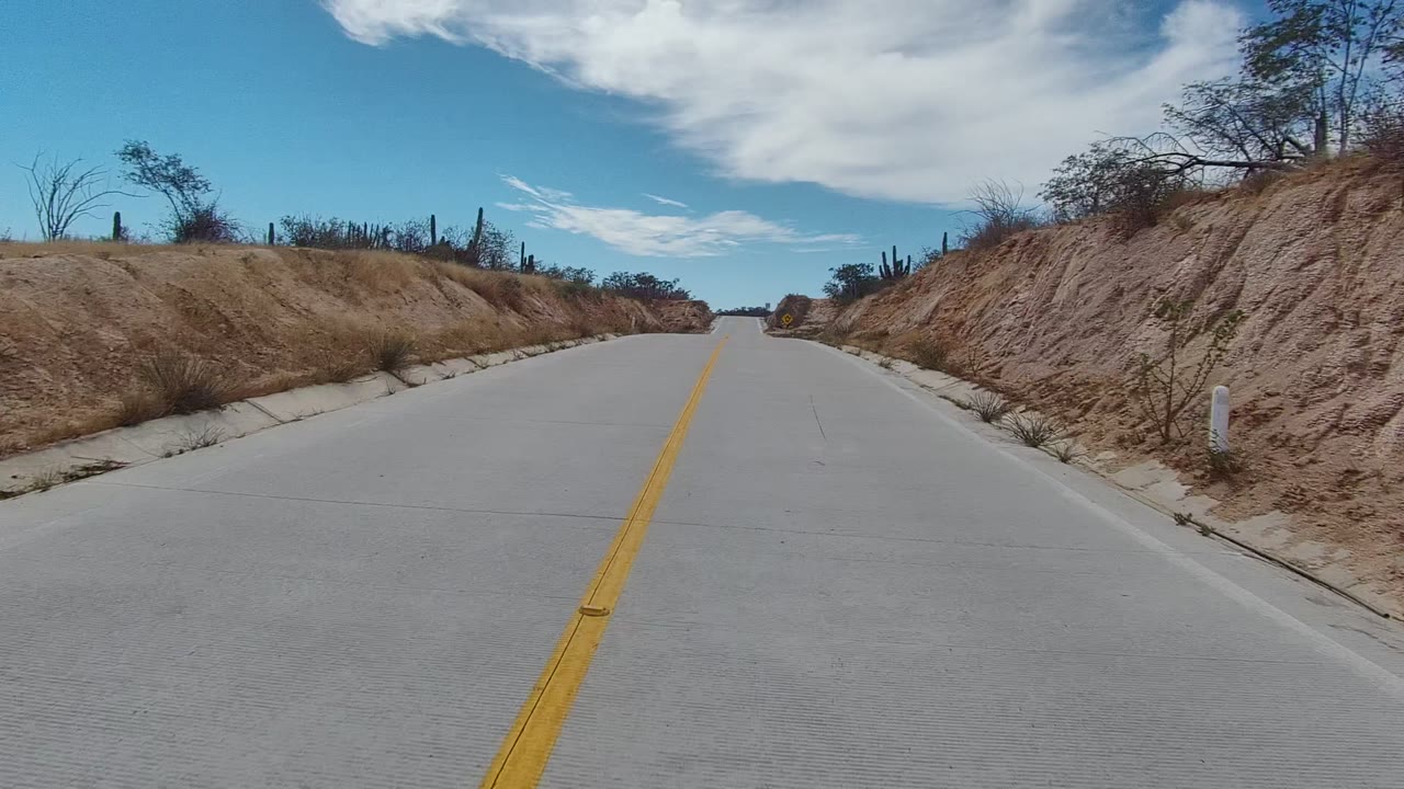 A straight road in East Cape Los Cabos runs between sandy cut banks with scattered cacti and scrub under a blue, clouded sky, with no visible people.