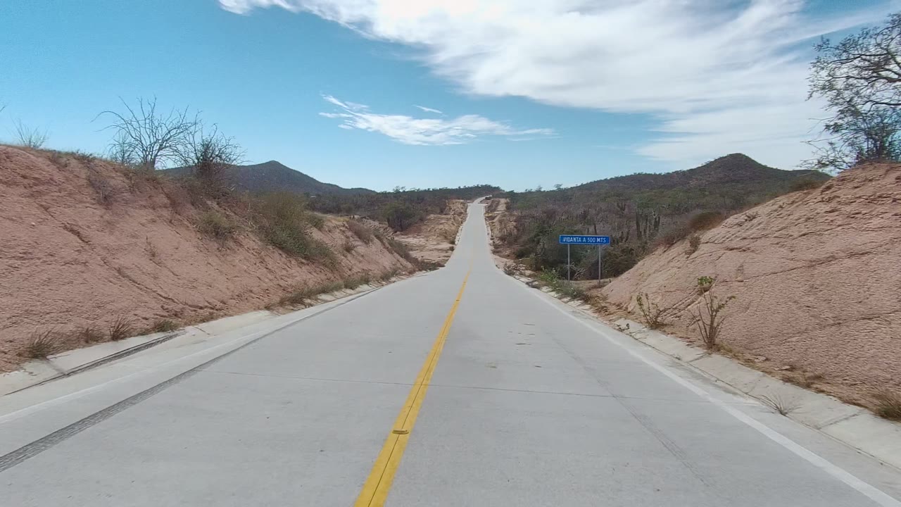 Opening frame of an East Cape Los Cabos road with a yellow center line, sandy roadside banks, distant hills, scattered clouds, and no visible people.
