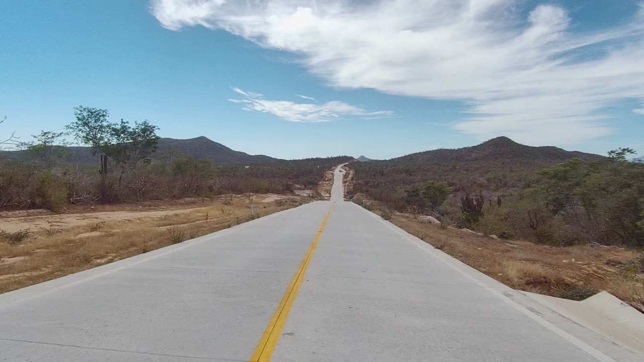 At 0:00, a straight concrete road in East Cape Los Cabos runs through dry roadside vegetation toward distant hills, with a yellow center line and no visible people.