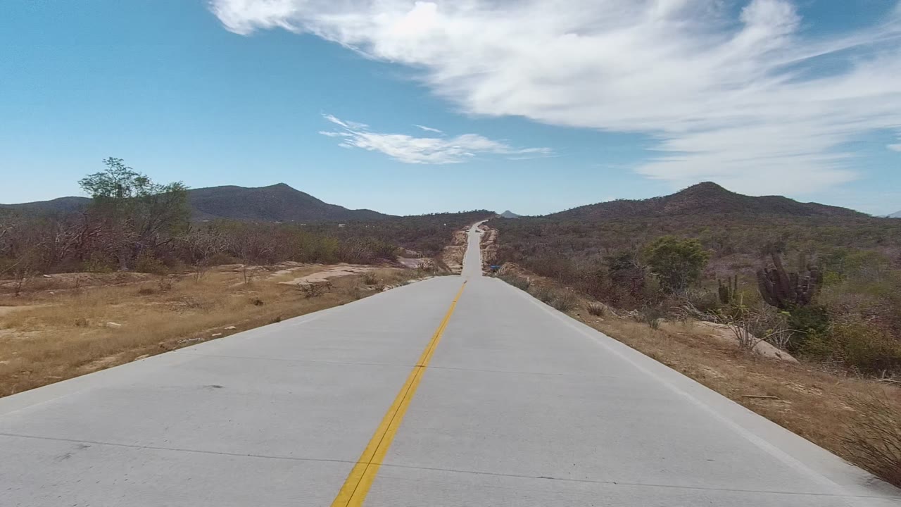 Roadway centered between dry brush and low hills with white clouds overhead.