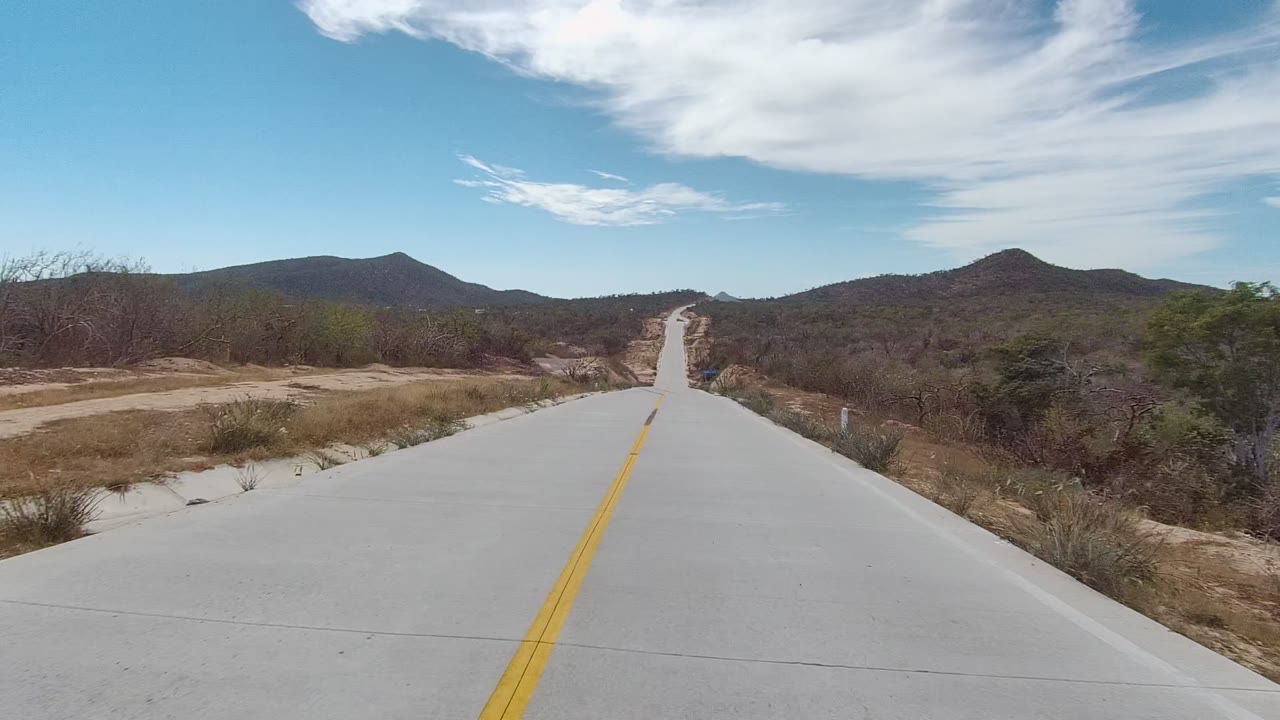 At 0:02, the road in East Cape Los Cabos passes sandy shoulders and sparse scrub, with low hills across the background and no visible people.