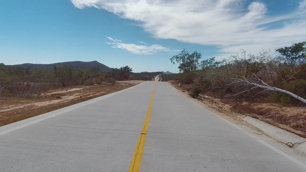 At 0:00, a paved East Cape Los Cabos road begins centered in frame, bordered by dry scrub, low trees, distant hills, blue sky, scattered clouds, and no visible people.