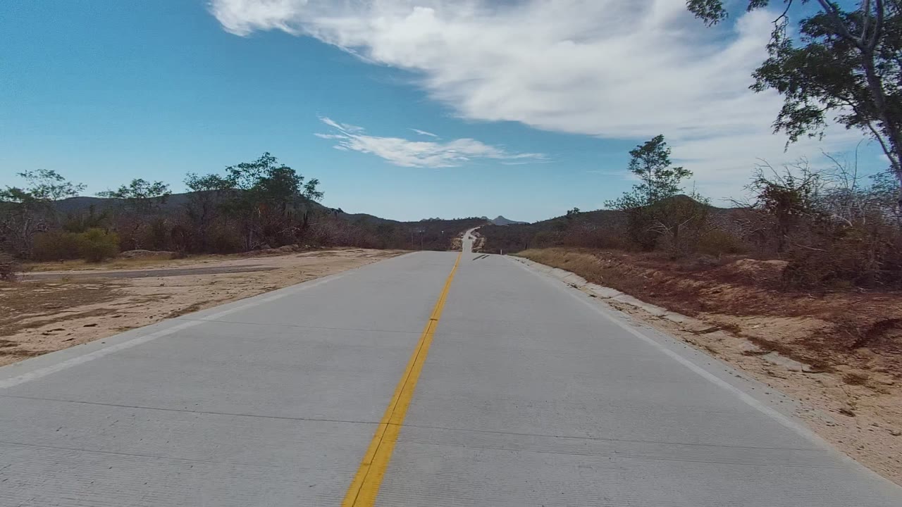 At 0:02, the road through East Cape Los Cabos bends slightly right, with sandy roadside ground, scrubby vegetation, low mountains, cloud streaks, and no visible people.