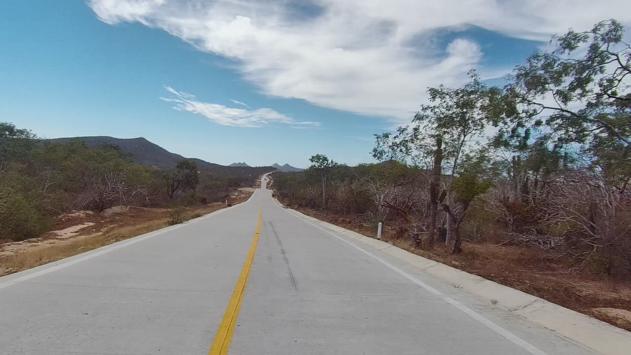 East Cape Los Cabos, Baja California Sur paved road centered between dry roadside brush, scattered trees, distant mountains, and bright clouds, with no visible people.
