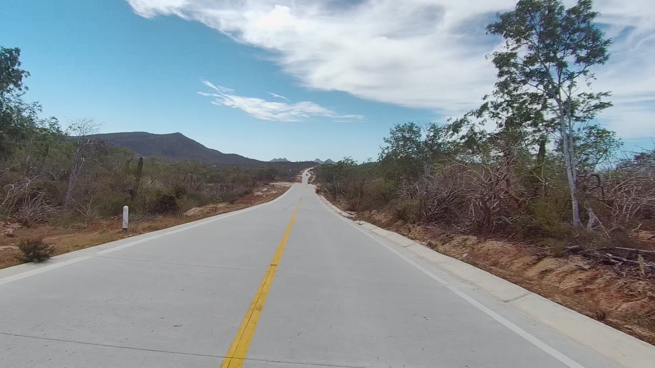 East Cape Los Cabos paved roadway running through sparse dry vegetation, with trees close to the right shoulder, mountains in the background, cloud bands overhead, and no visible people.