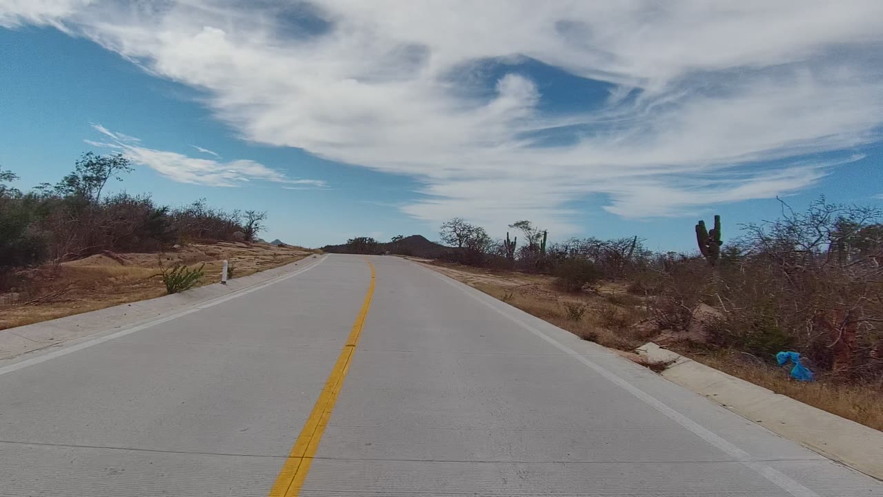 East Cape Los Cabos road view at the start of the clip, with the yellow center line leading between dry roadside brush, a cactus on the right, distant hills, and no visible people.