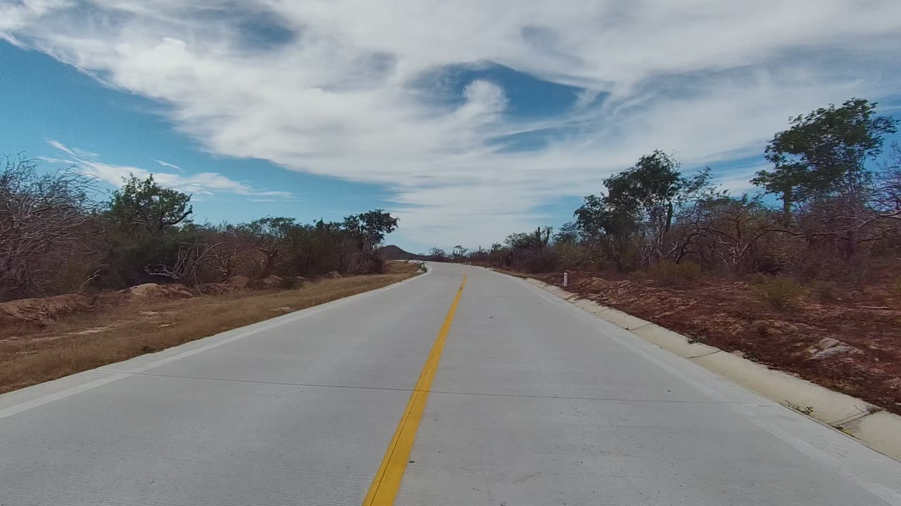 East Cape Los Cabos paved road opening into dry brush, scattered trees, low hills, blue sky, white clouds, and no visible people.