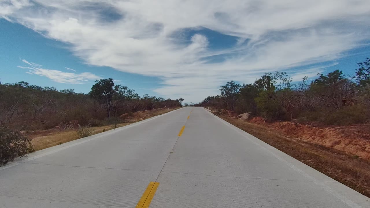 Start of a forward road view in East Cape Los Cabos, with pale pavement, a yellow center line, dry roadside shrubs, small trees, blue sky, white clouds, and no visible people.