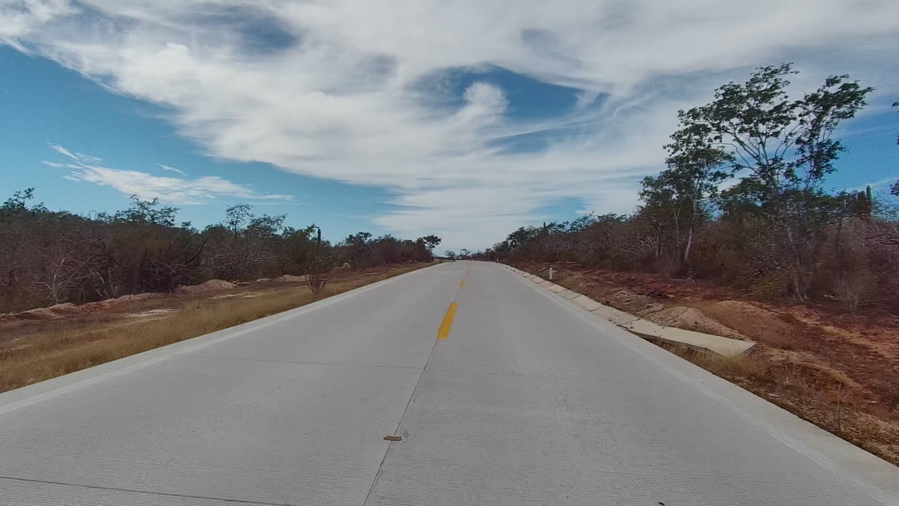 At 0:02, the East Cape Los Cabos roadway passes low brush and small trees, with a pale concrete edge visible on the right shoulder and no visible people.