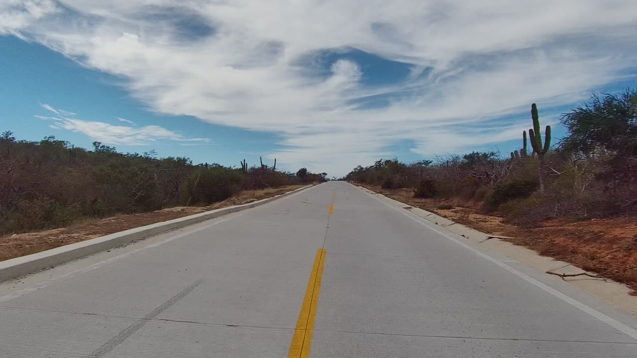 Forward view along a straight road in East Cape Los Cabos, Mexico, with a yellow center line, roadside cacti and dry vegetation, cloudy blue sky, and no visible people.