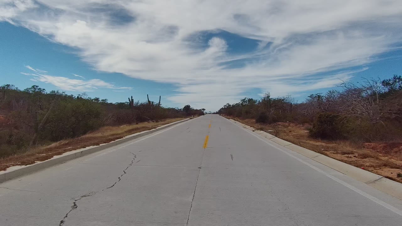 Empty road through dry brush with broad clouds overhead.