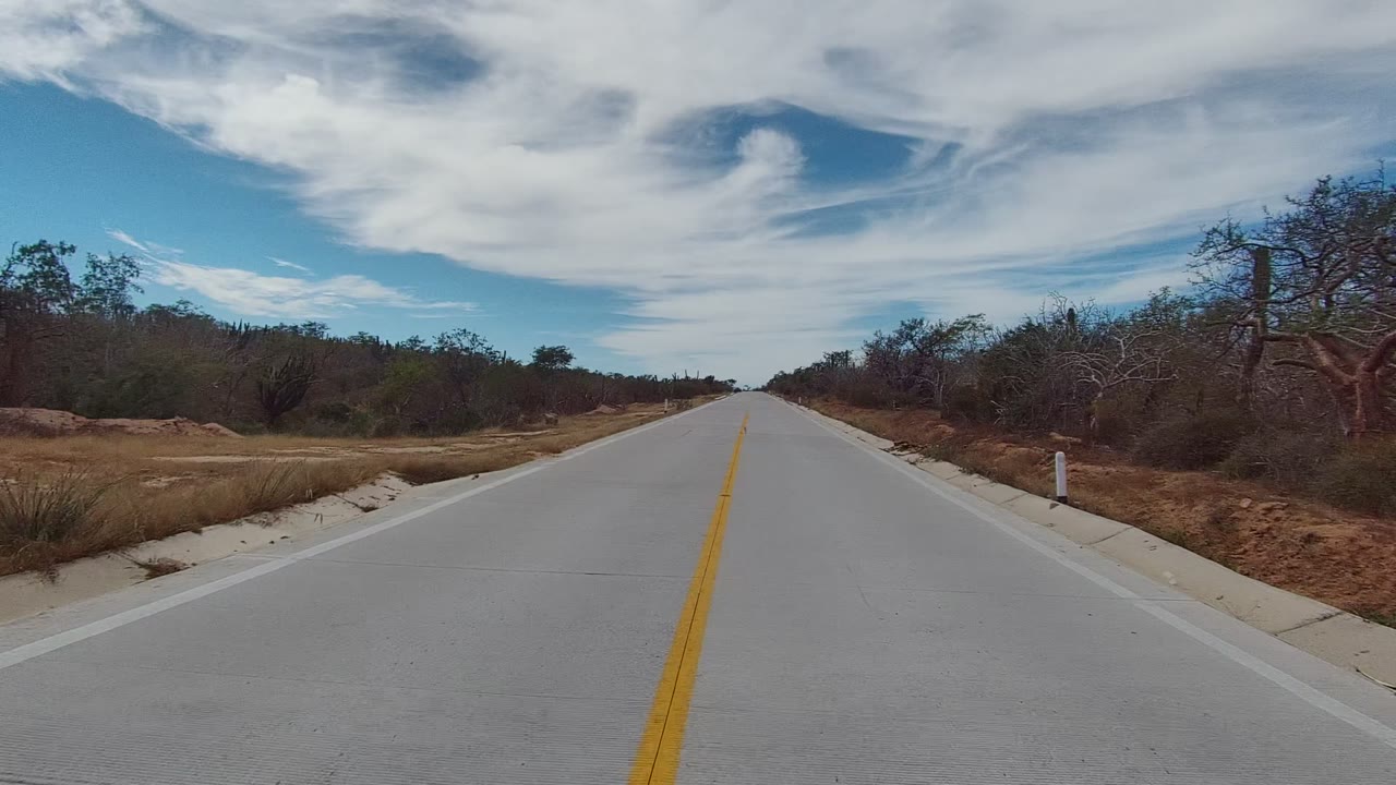 Straight paved road in East Cape Los Cabos, Baja California Sur, with a yellow center line, dry roadside brush, low trees, and no visible people.