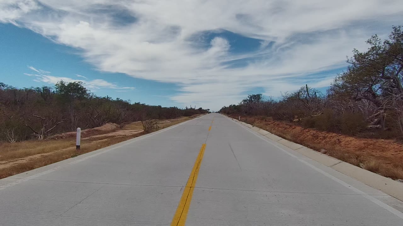 Forward road view in East Cape Los Cabos with the yellow line shifting toward the right foreground, reddish roadside soil, dry shrubs, and no visible people.