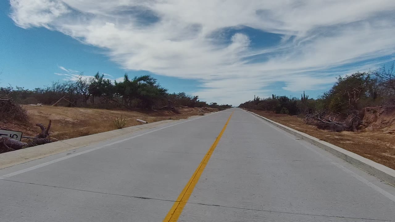 Start of the East Cape Los Cabos road clip, showing a straight pale roadway with a yellow center line, dry shoulders, low bushes, distant cacti, blue sky, and no visible people.