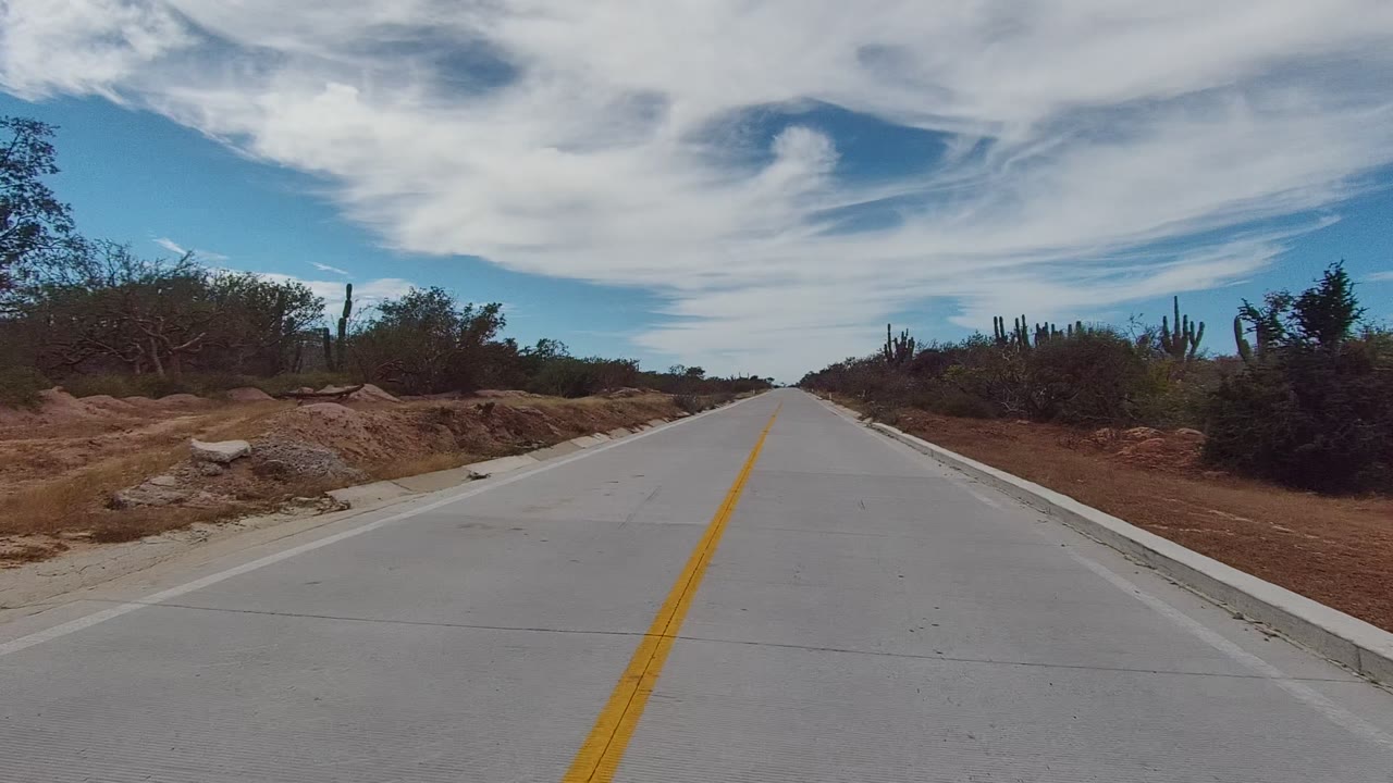 At 0:02, the East Cape Los Cabos roadway continues ahead with a yellow stripe, exposed dry soil, scattered bushes, several cactus forms, and no visible people.