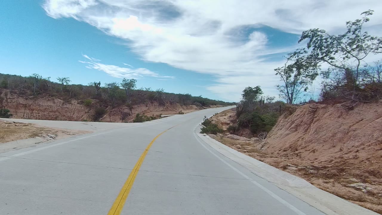 Start of the East Cape Los Cabos road view, with a pale paved lane, yellow center line, sandy roadside banks, sparse trees, and no visible people.