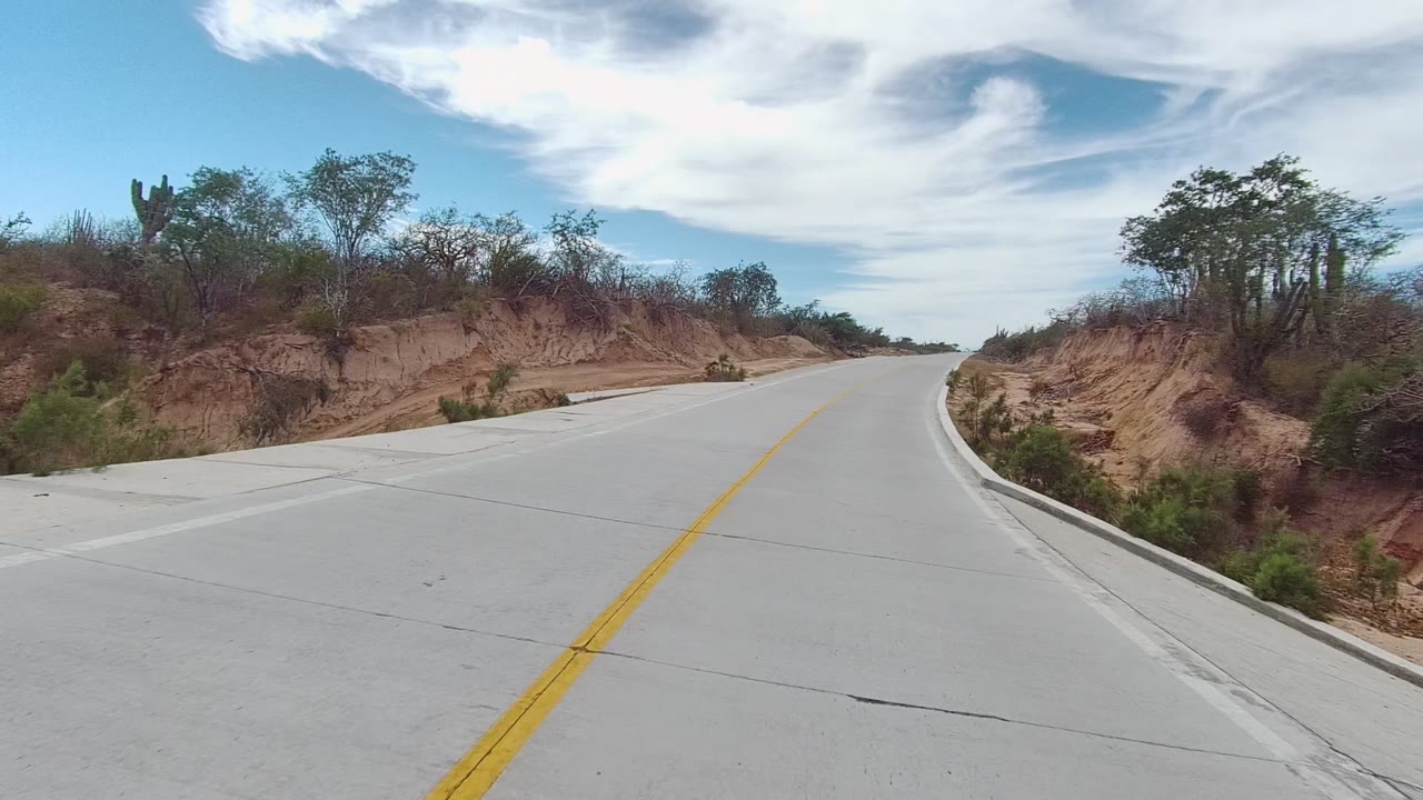 A later East Cape Los Cabos frame shows the paved road bending right, with cactus shapes and scrub along the sandy slopes, bright sky above, and no visible people.