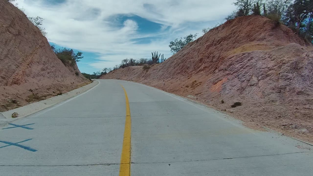 Opening frame of a paved road in East Cape Los Cabos, Mexico, with a yellow center line, reddish dirt banks, sparse brush, cacti, and no visible people.
