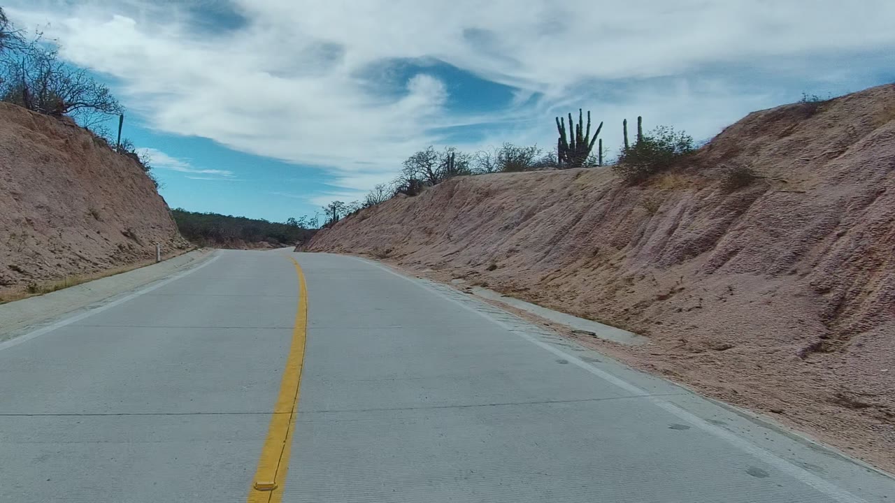 The road passes close to a reddish dirt embankment in East Cape Los Cabos, Mexico, with cacti and dry brush on the ridge and no visible people.