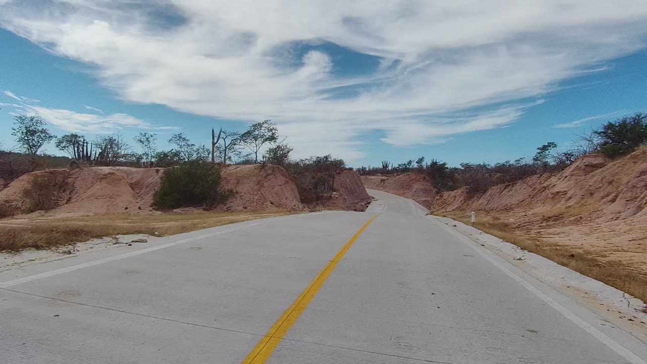 A paved road with a yellow center line runs into red-brown arid hills in East Cape Los Cabos, Mexico, with sparse brush and no visible people.