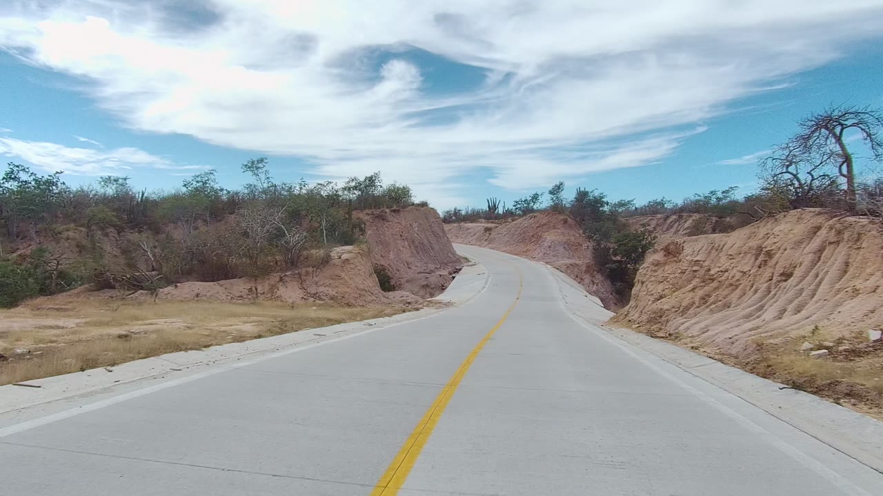 A pale concrete road arcs past sandy red-brown banks, low brush, and cactus-like plants in East Cape Los Cabos, Mexico, with no visible people.