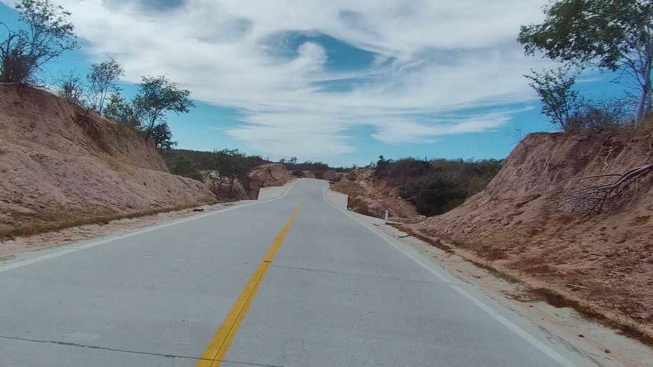 A paved road in East Cape Los Cabos, Baja California Sur begins between dry cut banks and sparse roadside trees under streaked clouds, with no visible people.
