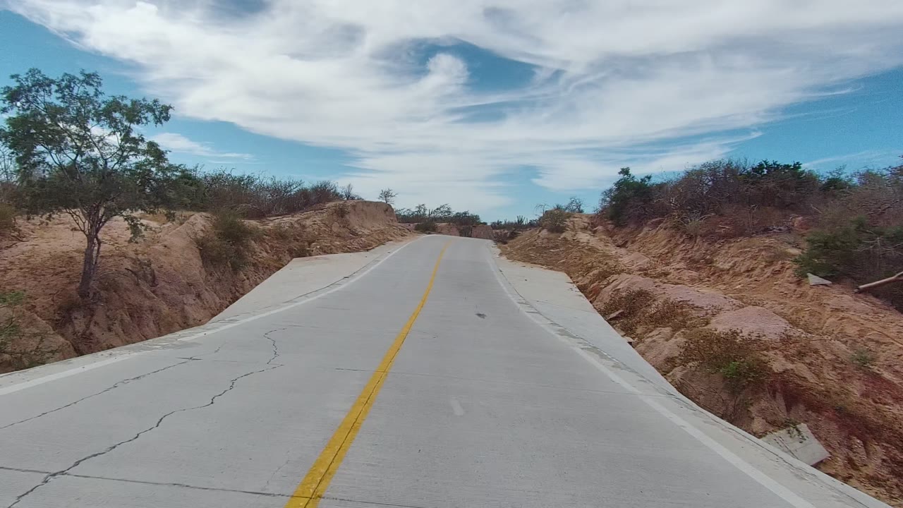 A closer view of the paved route in East Cape Los Cabos, Baja California Sur passes cracked concrete, eroded roadside banks, scattered trees, and no visible people.