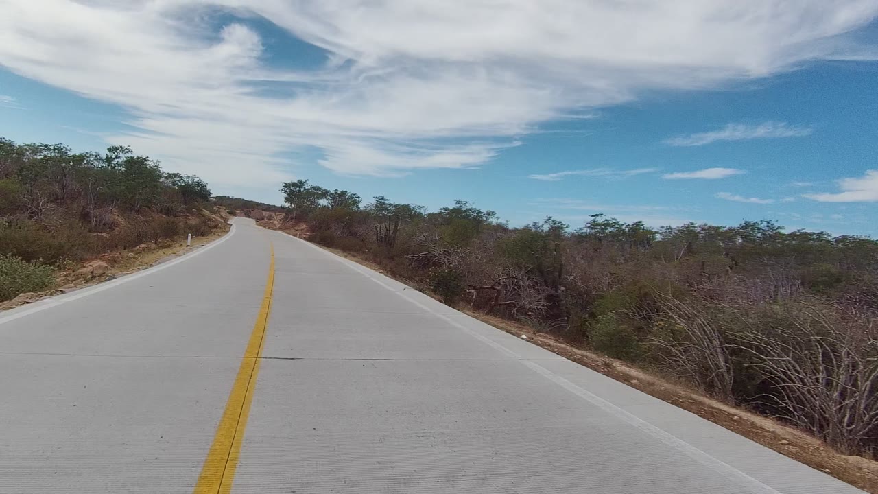 Opening view of an empty paved road in East Cape Los Cabos, Baja California Sur, with dry roadside brush, a yellow center line, blue sky, white clouds, and no visible people.