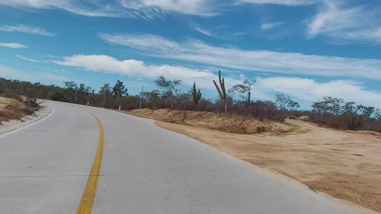 Vehicle-height view of a pale road curving left through dry East Cape Los Cabos scrub, with cacti, sandy shoulder, scattered clouds, and no visible people.