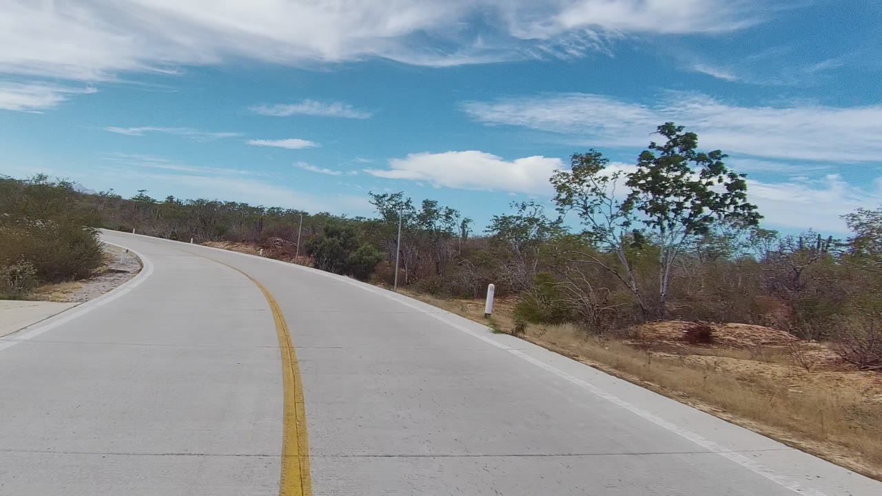 An empty paved road in East Cape Los Cabos curves left past scrubby vegetation, sandy ground, white roadside markers, and a bright clouded sky, with no visible people.