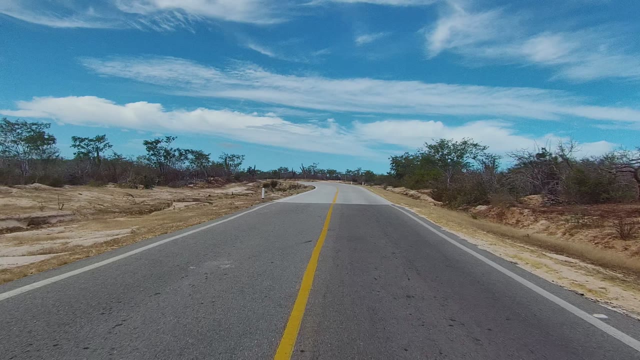 At 0:00, a paved road in East Cape Los Cabos runs straight ahead before curving through dry sandy ground with low brush, small trees, blue sky, and no visible people.