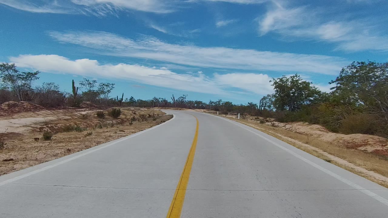 At 0:02, the road curve opens wider in East Cape Los Cabos, with pale pavement, a yellow center line, dry brush, cactus silhouettes, sandy banks, and no visible people.