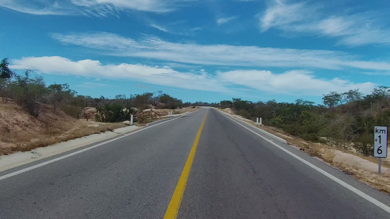 Straight road in East Cape Los Cabos, Baja California Sur, with a yellow center line, white edge posts, dry scrub on both sides, scattered clouds, and no visible people.