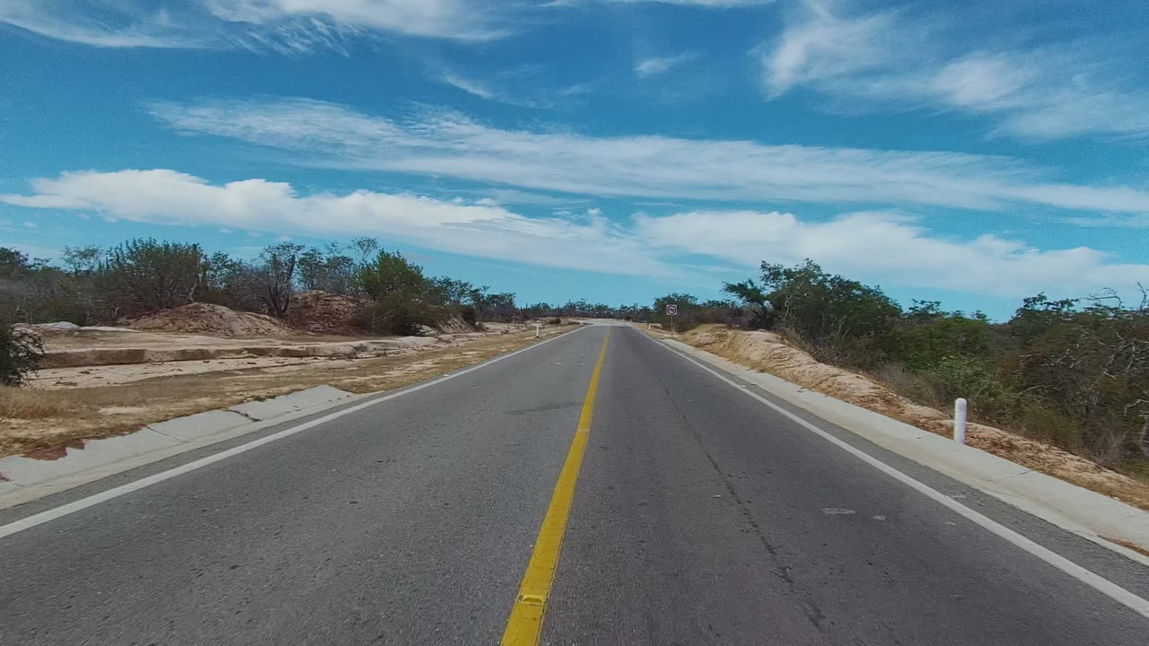 The road remains centered in East Cape Los Cabos, with a long yellow stripe, pale sandy shoulders, scrubby vegetation, a low horizon, bright blue sky, and no visible people.