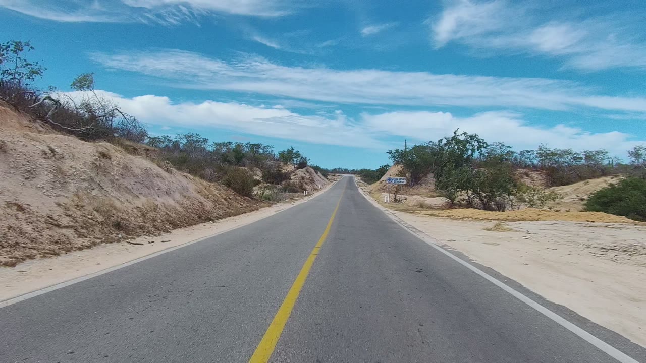 A centered paved road in East Cape Los Cabos, Mexico runs between sandy banks and sparse trees under a blue sky, with no visible people.