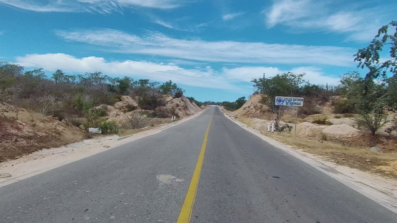 Paved road passing dry scrub and a roadside sign in sandy terrain.