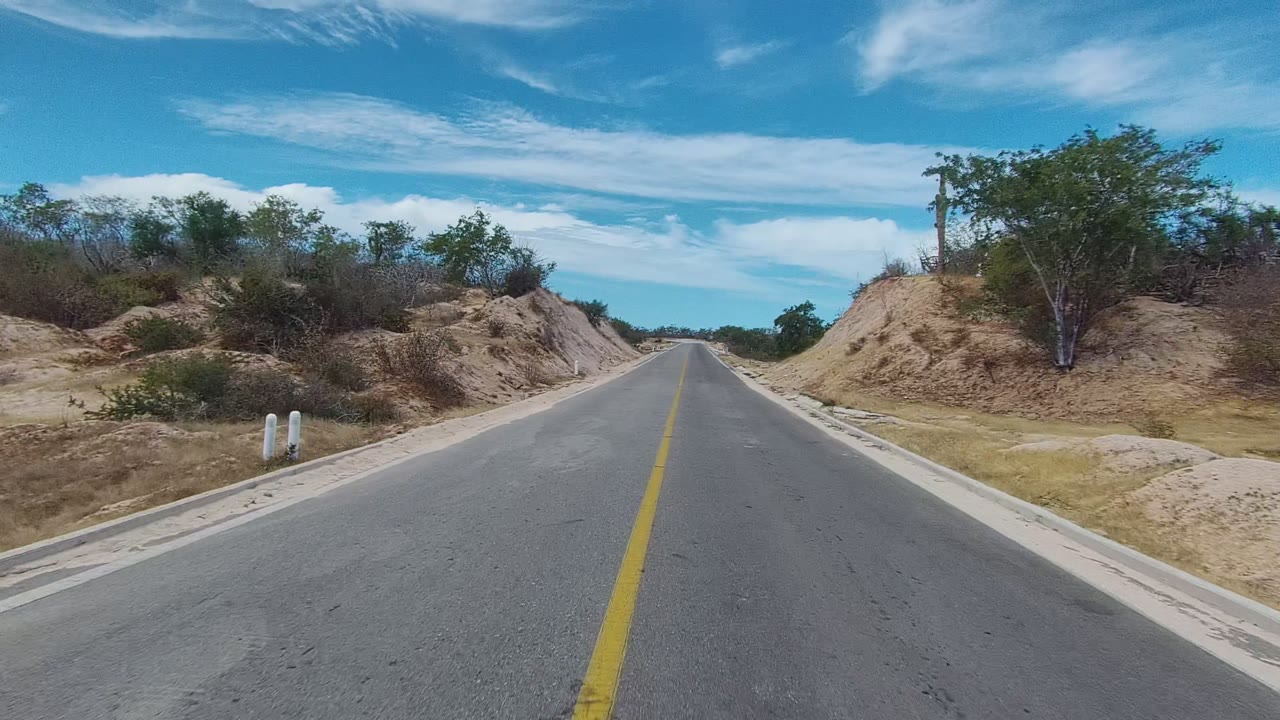 An empty paved road in East Cape Los Cabos, Mexico stretches past sandy banks, white roadside posts, scrubby trees, and bright clouded sky, with no visible people.