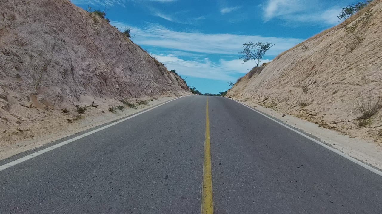 Paved East Cape Los Cabos road begins between steep sandy hills, with a yellow center line, sparse scrub, blue sky, and no visible people.