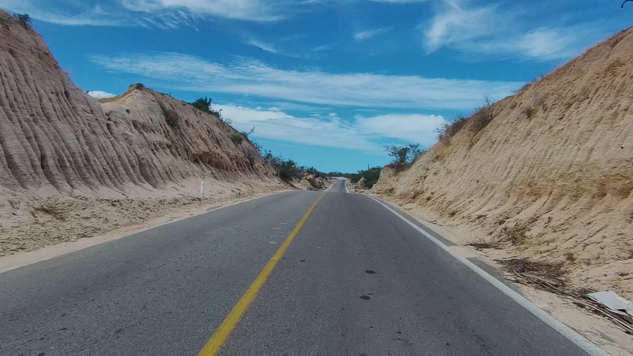 A straight paved road in East Cape Los Cabos passes a ribbed sandy mound on the right, with dry brush and open blue sky, and no visible people.