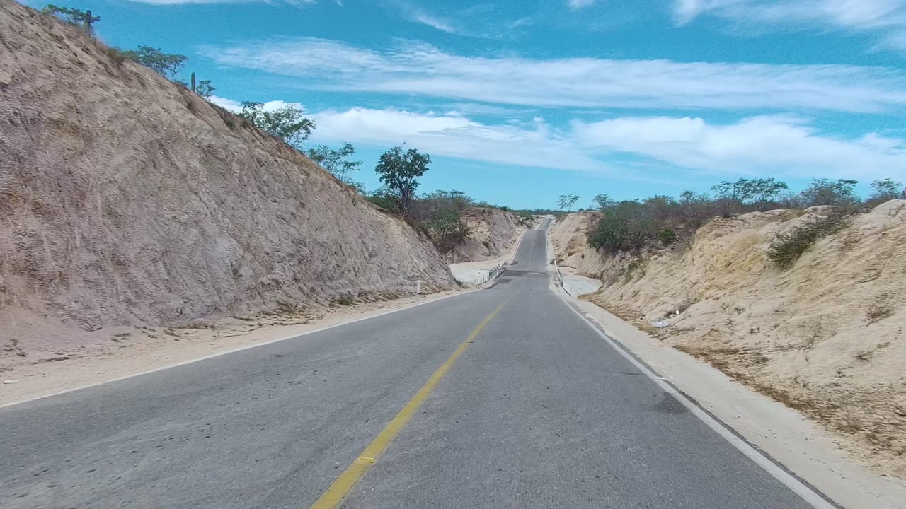 Paved road in East Cape Los Cabos, Baja California Sur, running between dry rocky slopes with sparse shrubs and no visible people.