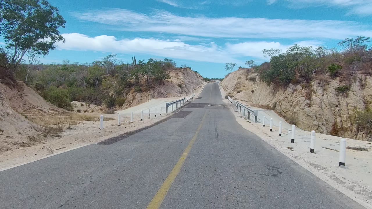 Empty paved road in East Cape Los Cabos, Baja California Sur, bordered by white posts, metal guardrails, eroded sandy banks, sparse vegetation, and no visible people.