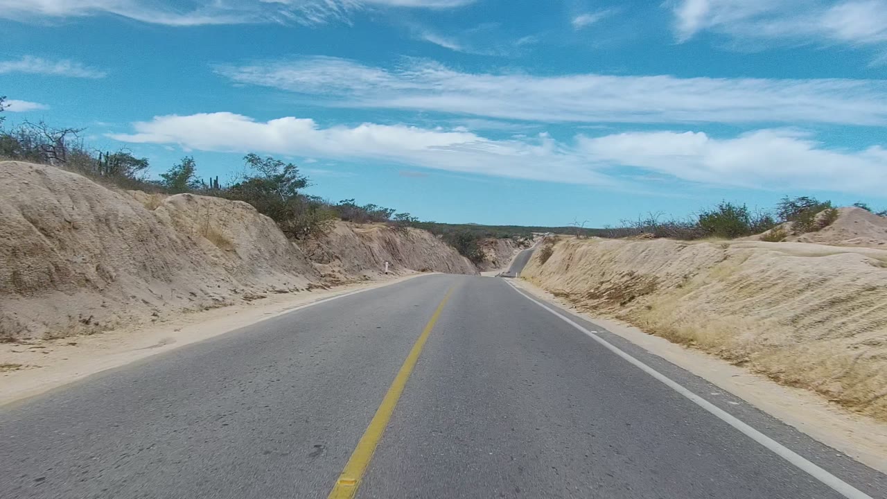 Straight paved road in East Cape Los Cabos, Baja California Sur, narrowing between sandy cuts with a cactus on the left side, low shrubs, bright clouds, and no visible people.